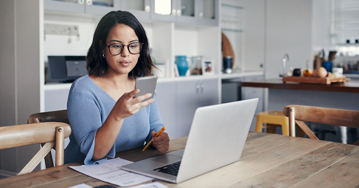 woman sitting at kitchen table going through finances on phone and computer Thumbnail